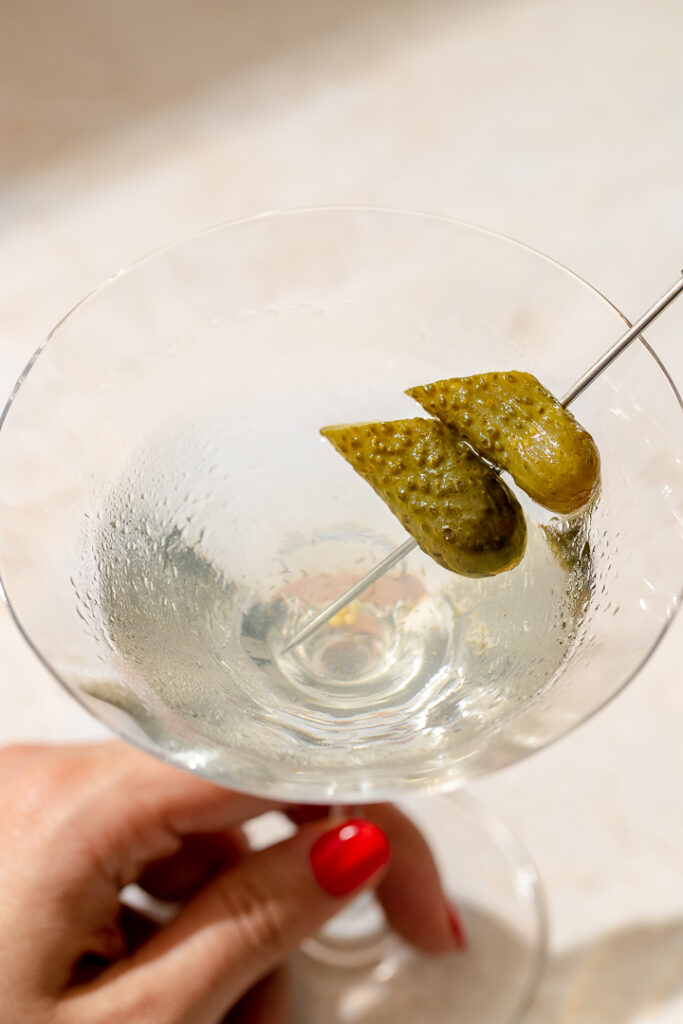A hand holding a pickle martini with a gherkin as a garnish