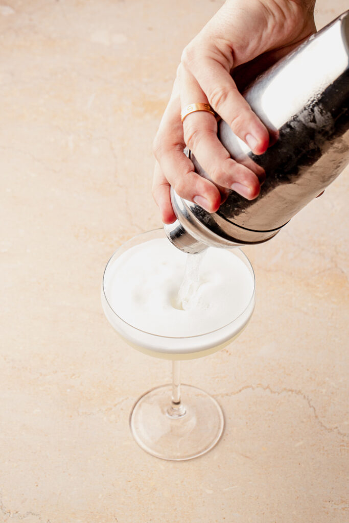 A silver shaker pouring cocktail into a coupe glass