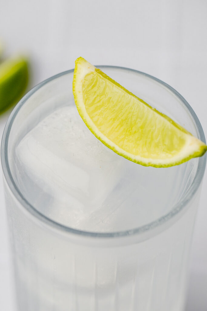Close up shot of the top of a collins glass with a clear liquid inside, ice and a lime wedge