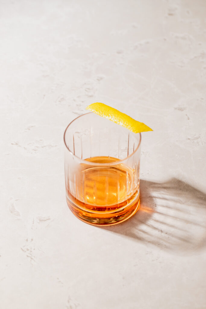 A rocks glass with amber liquid in the bottom and a garnish of lemon zest sitting on the rim.