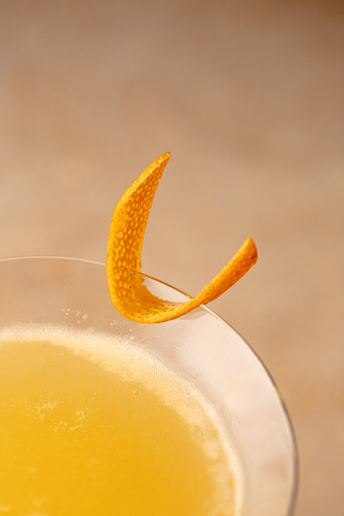 Close up of orange zest garnish sitting on the rim of a martini glass filled with an amber liquid.