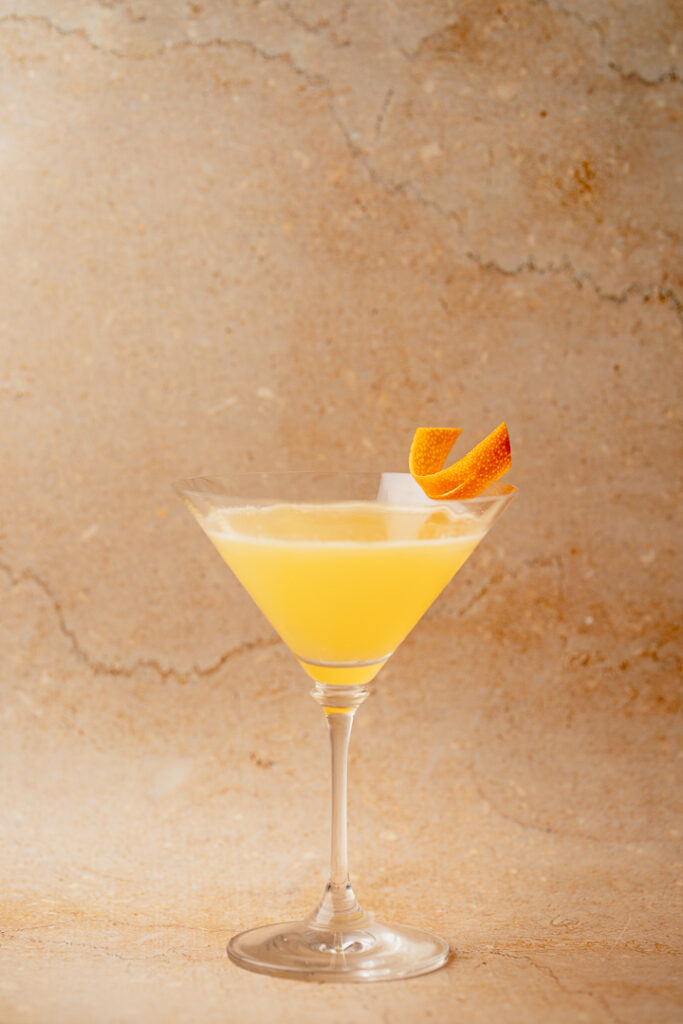 A martini glass with a yellow orange liquid inside and orange zest on the rim
