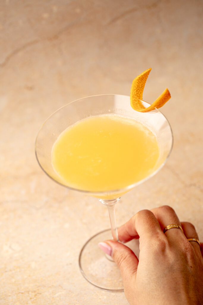 A hand holding the stem of a martini glass with a yellow orange liquid inside and orange zest on the rim