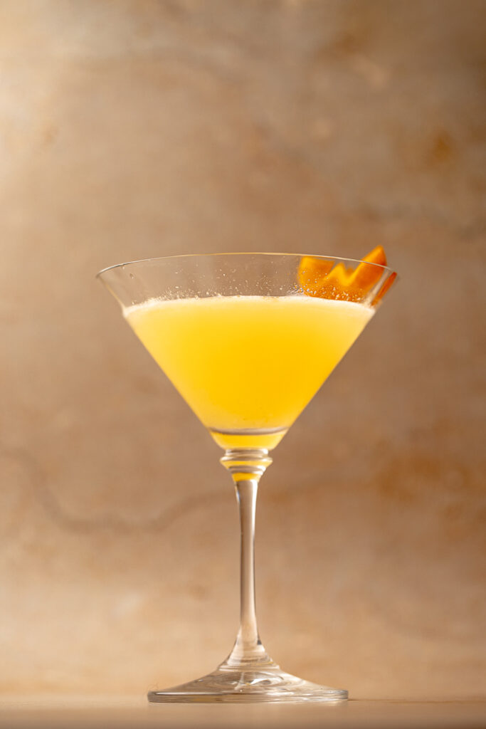 A martini glass with a yellow orange liquid inside and orange zest on the rim