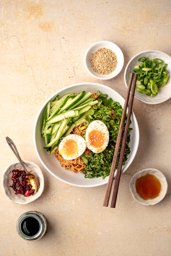 A top down shot of a bowl of cold peanut noodles garnished with cucumber, spring onion, coriander and two halves of a soft boiled egg. Surrounded by small bowls of different condiments.