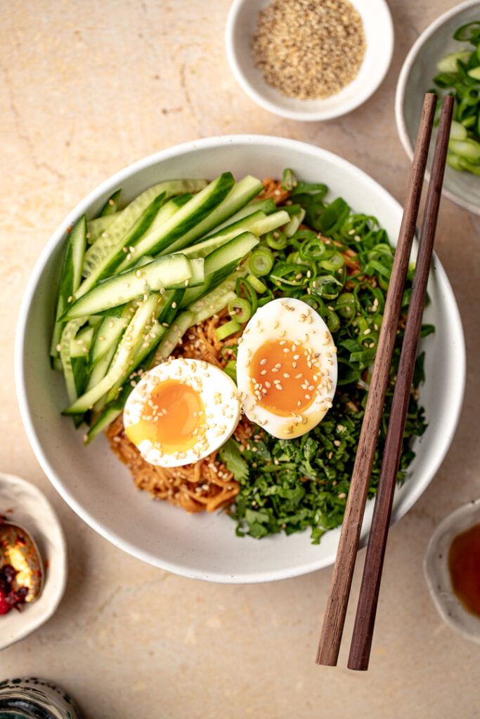 A top down shot of a bowl of cold peanut noodles garnished with cucumber, spring onion, coriander and two halves of a soft boiled egg. 