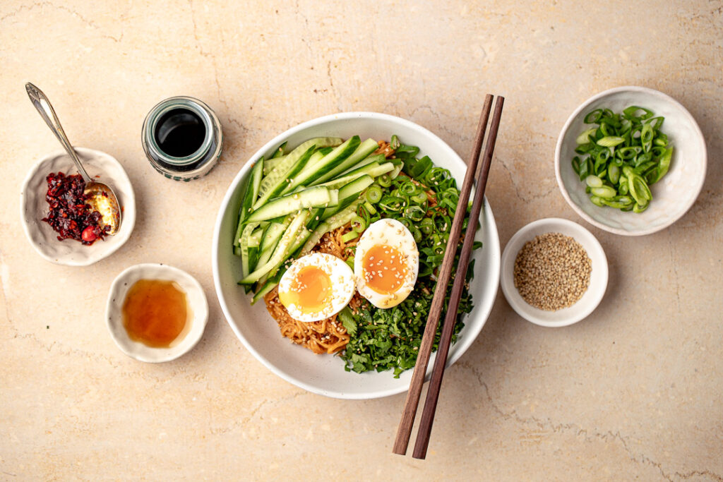 A top down shot of a bowl of cold peanut noodles garnished with cucumber, spring onion, coriander and two halves of a soft boiled egg. Surrounded by small bowls of different condiments.