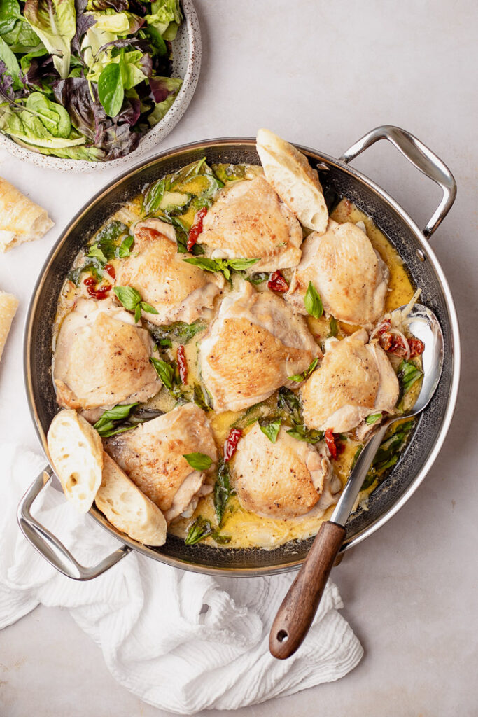 A pan with a serving spoon resting on the side with chicken in a creamy sauce with sun dried tomatoes and basil leaves. A plate of dressed green salad leaves sits above the pan and slices of crusty bread rest on the side.