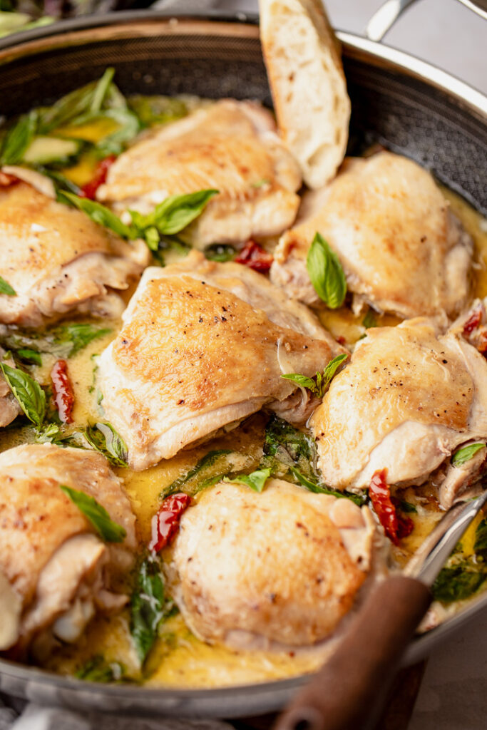 A pan filled with pieces of chicken in a creamy sauce dotted with sun dried tomatoes and basil leaves. 