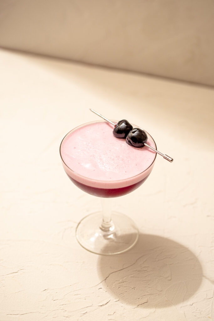 The top of a coupe glass with a dark pink-purple liquid, a light pink foam on top. Topped with two maraschino cherries on a cocktail pick.