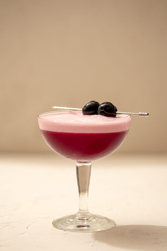 A side on shot of a coupe glass with a dark pink-purple liquid, a light pink foam on top. Topped with two maraschino cherries on a cocktail pick.