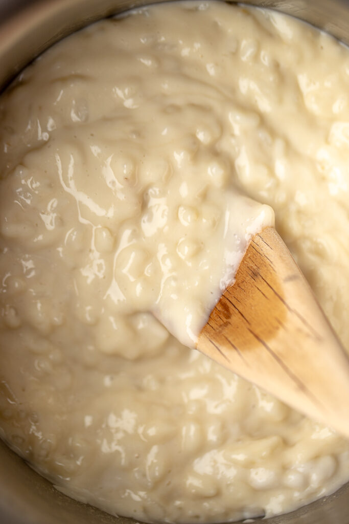 A close up, top down shot of creamy coconut rice pudding in a pot with a wooden spoon resting in it.