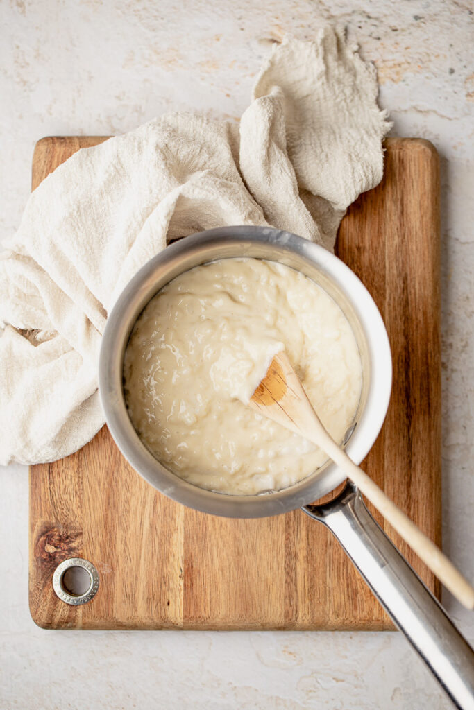 Creamy coconut rice pudding in a pot with a wooden spoon resting in it, sitting atop a chopping board.