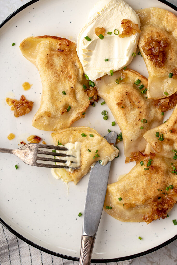 A plate of fried pierogi ruskie with sour cream, sprinkled with sautéed onions and chives