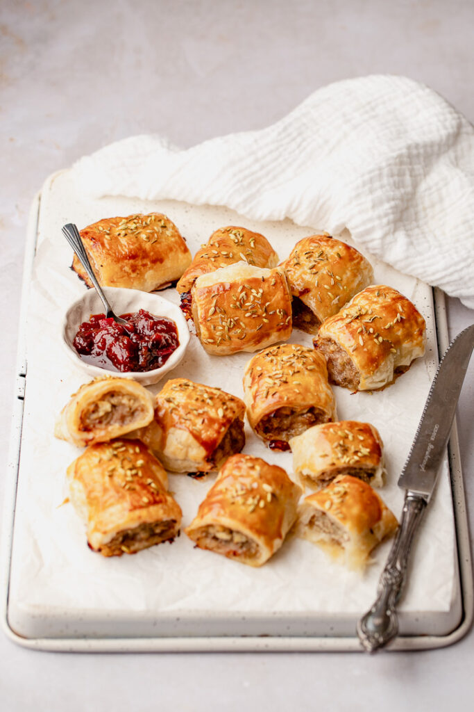 Pork and apple sausage rolls on a white tray with a silver knife resting on the edge, with a small bowl of tomato relish.