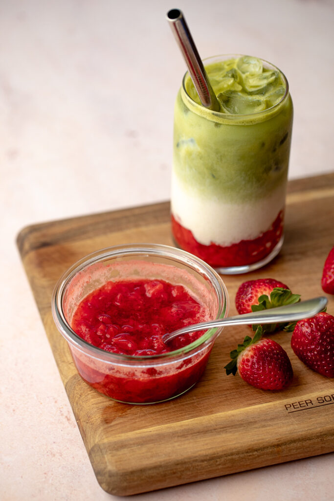 Iced strawberry matcha latte, small glass container of strawberry compote and fresh strawberries sitting atop a wooden board.