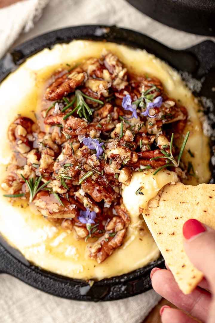 Baked brie with pecans, honey and rosemary in a black dish with a cracker being dipped in.