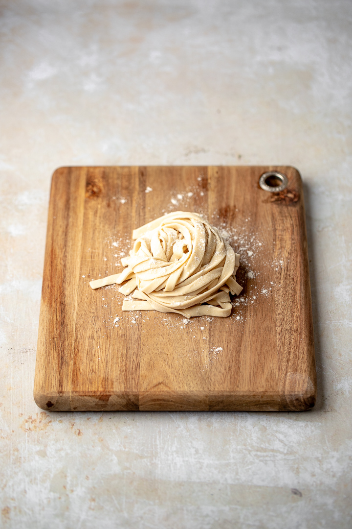 A wooden board with a nest of floured knife cut noodles on top.