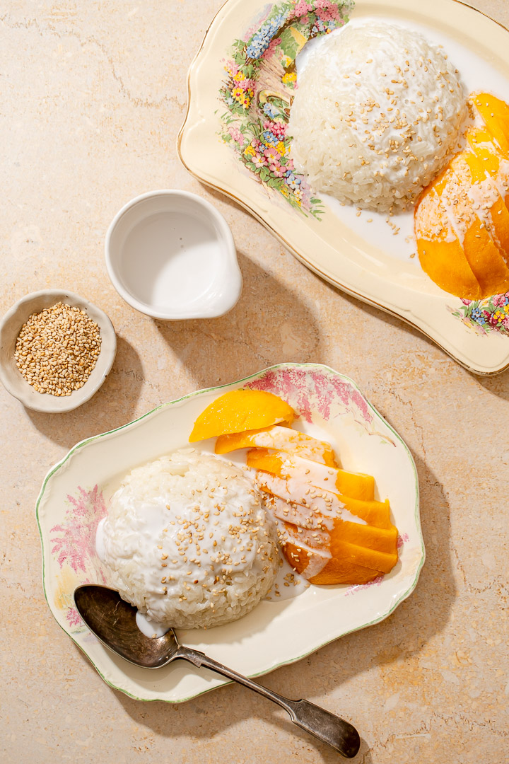 Two rectangular plates with glutinous rice and fresh sliced mango, drizzled in coconut sauce and sprinkled with toasted sesame seeds. 