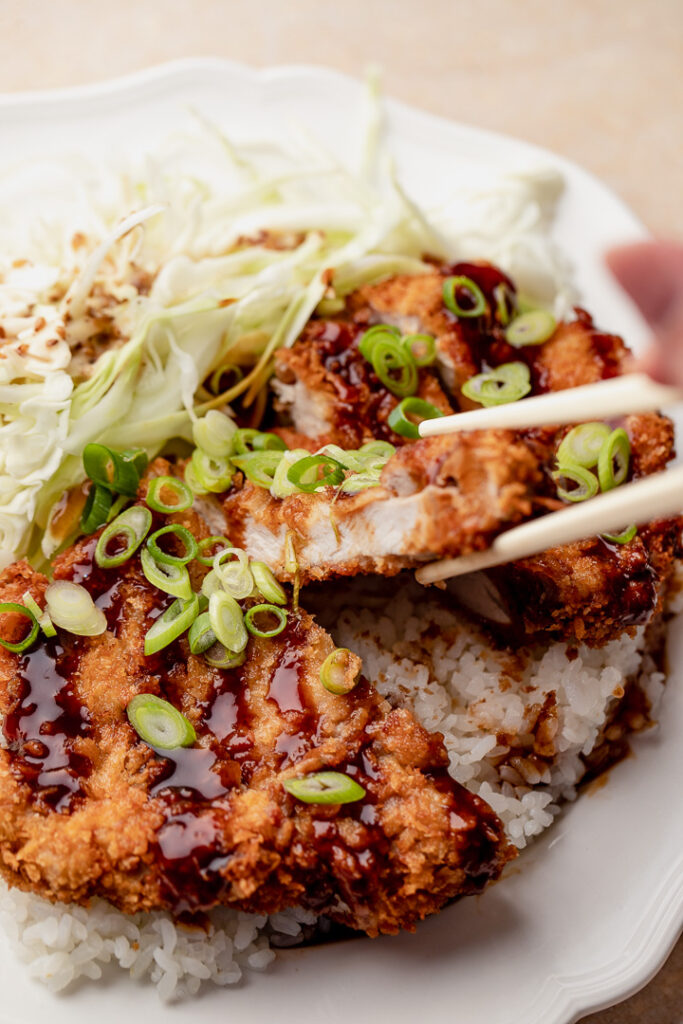 Chopsticks lifting a slice of pork tonkatsu on a bed of rice with cabbage salad on the side.