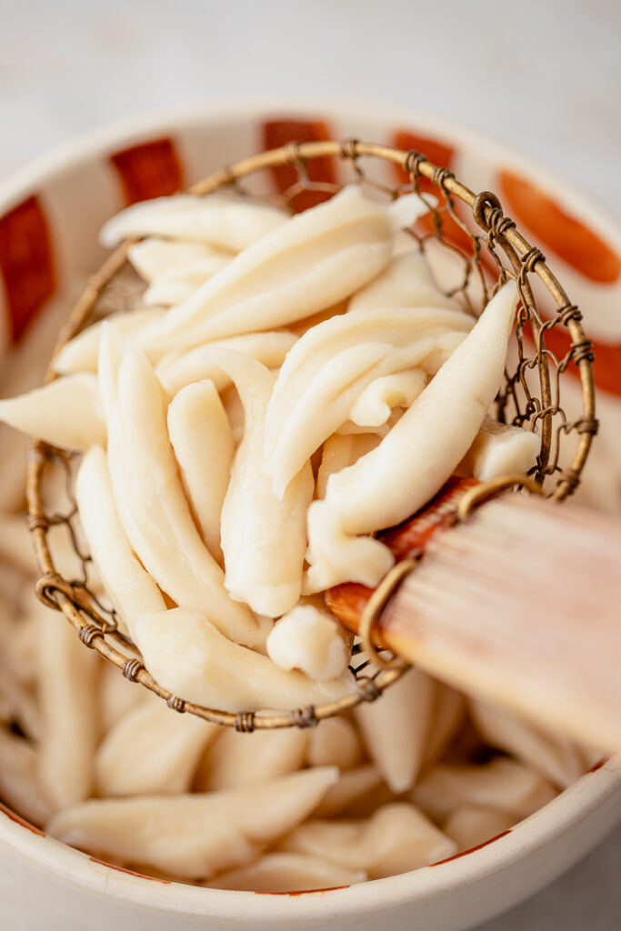 A wire skimmer scooping up cooked scissor-cut noodles from a bowl