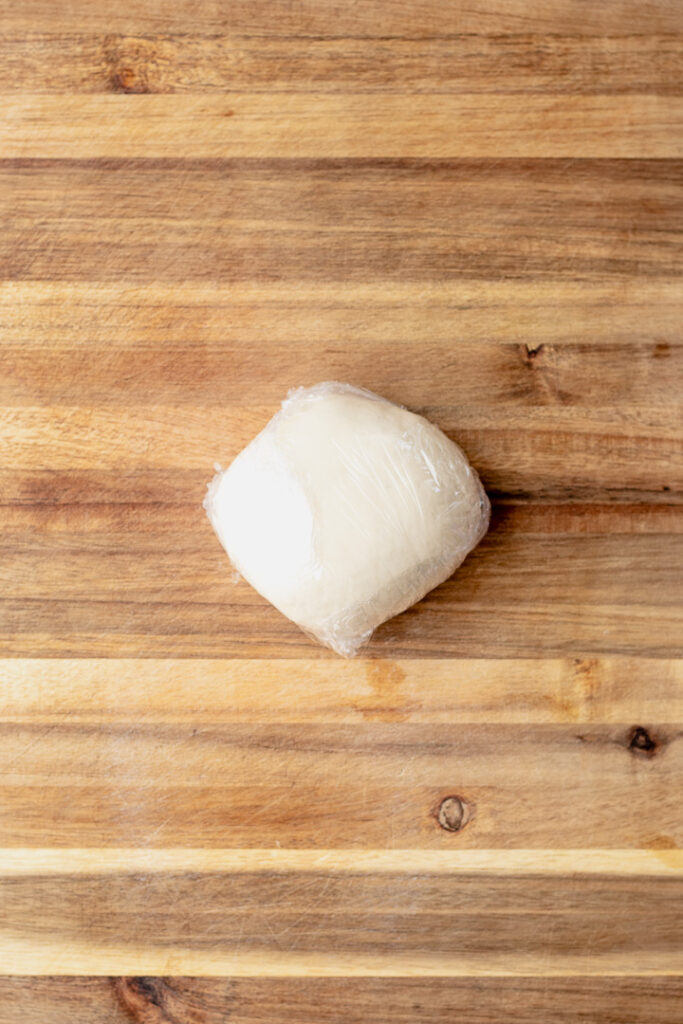 A ball of dough covered in plastic wrap sitting on a wooden chopping board